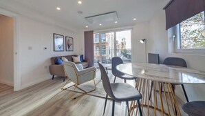 A modern living room with a round glass table, grey chairs, a sofa with cushions, and a large window offering a view of buildings. - 2BR Apartment near Ashford Town | Balcony & WiFi (Ashford)