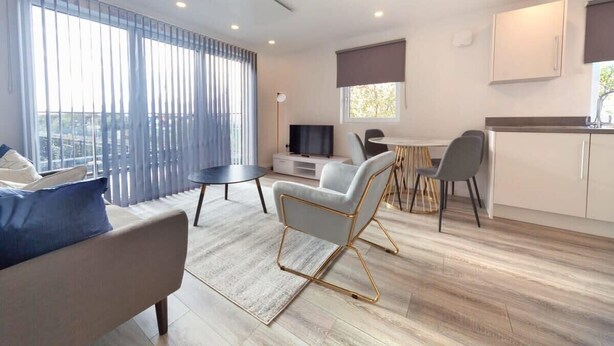A modern living room with a sofa, a dining table, and chairs, a kitchen area, and a large window with curtains. - 2BR Apartment near Ashford Town | Balcony & WiFi (Ashford)