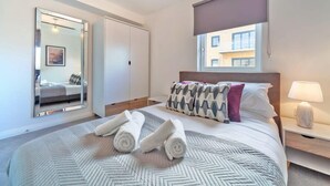 A modern hotel room with a large bed, a bedside table with a lamp, a window with a view of buildings, and a wardrobe. - 2BR Apartment near Ashford Town | Balcony & WiFi (Ashford)
