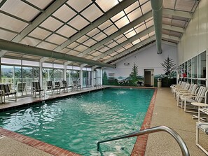 An indoor swimming pool with clear water, a tiled edge, and a metal ladder. There are lounge chairs and a view of the outdoors through large windows. - Whispering Pines 124 by Jackson Mountain Rentals (Pigeon Forge)