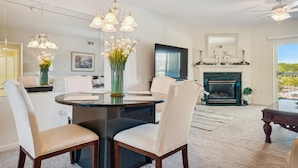 A dining area with a round table, chairs, and a chandelier. There is a fireplace with a mantel and a TV mounted on the wall.