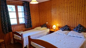 Een kamer met houten lambrisering, twee enkele bedden, een stoel, een tafel met een lamp en een raam met gordijnen.