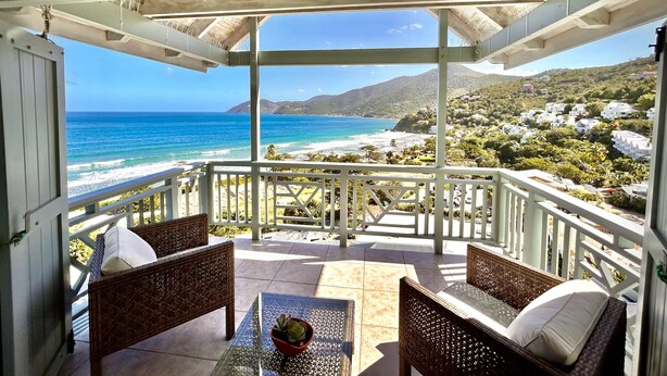 Un espace extérieur aménagé, abrité et doté de mobilier en osier et d’une petite table, offrant une vue sur une plage et une ville située sur une colline.