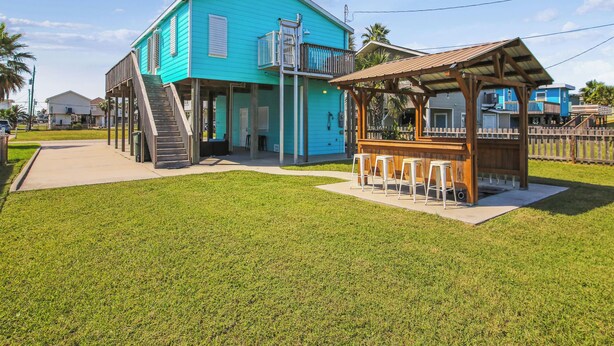 A turquoise two-story building with a covered outdoor seating area and a staircase leading to a balcony. - The Wooden Fin by Ryson Vacation Rentals (Galveston)
