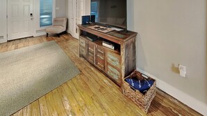 A room with wooden flooring, a large TV, a wooden cabinet, a wicker basket, and a bench. - The Wooden Fin by Ryson Vacation Rentals (Galveston)