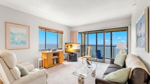 A spacious living room with a large TV, comfortable seating, a coffee table, and a balcony with a view of the sea. - Ocean Sands 1101 by Distinctive Beach Rentals (Madeira Beach)