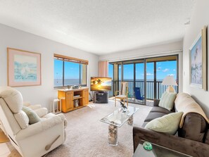 A spacious living room with a large TV, comfortable seating, a coffee table, and a balcony with a view of the sea. - Ocean Sands 1101 by Distinctive Beach Rentals (Madeira Beach)
