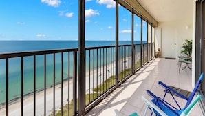A balcony with a view of the ocean and beach, featuring a door, a potted plant, and two blue chairs. - Ocean Sands 1101 by Distinctive Beach Rentals (Madeira Beach)