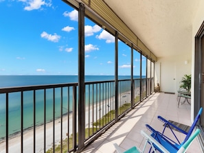 A balcony with a view of the ocean and beach, featuring a door, a potted plant, and two blue chairs. - Ocean Sands 1101 by Distinctive Beach Rentals (Madeira Beach)
