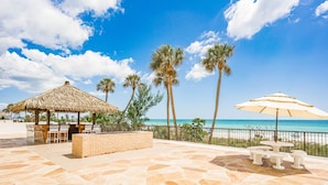 A beachside patio with a thatched-roof structure, a seating area with benches, and a view of the ocean. - Ocean Sands 1101 by Distinctive Beach Rentals (Madeira Beach)