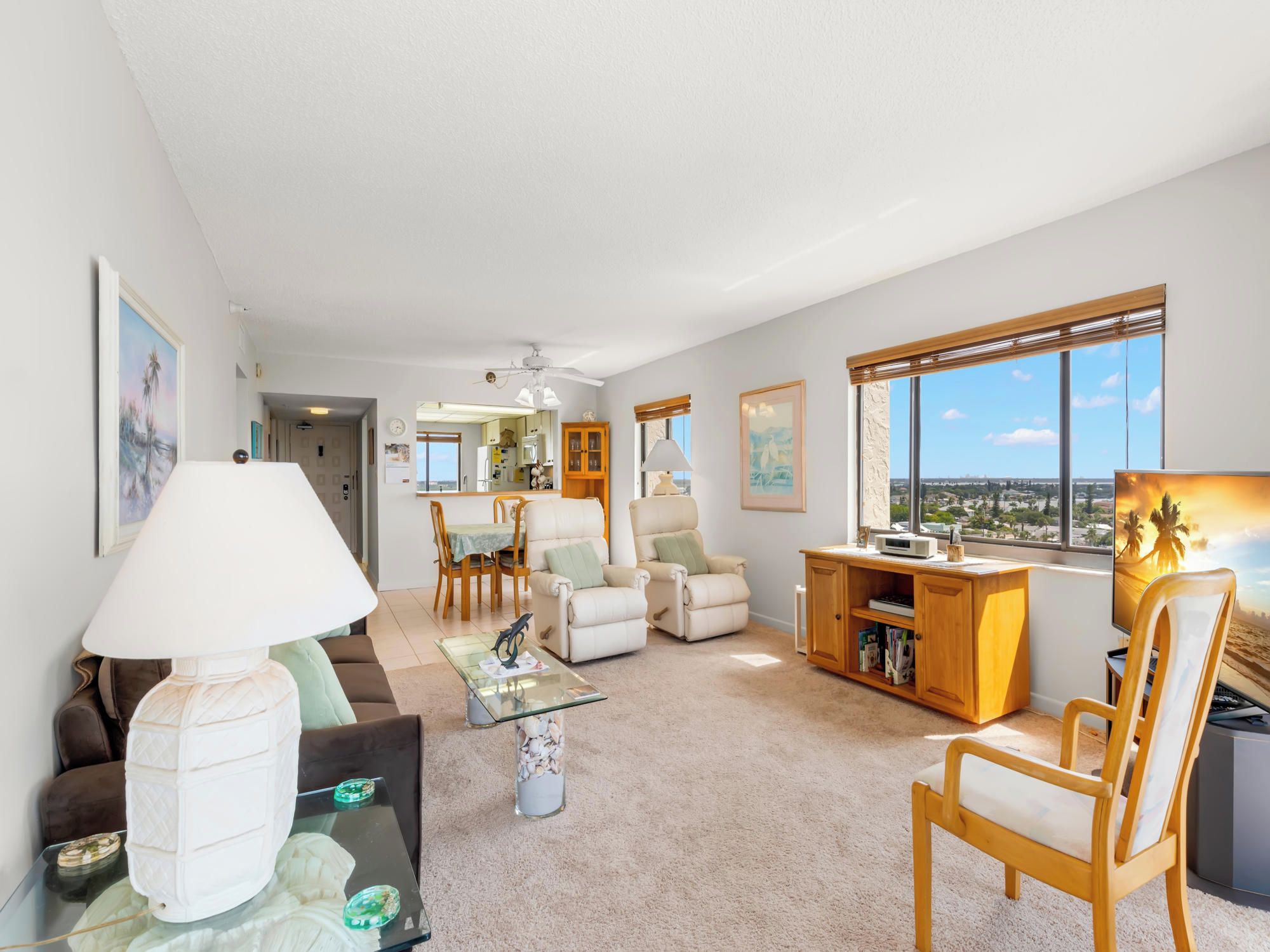 A spacious living room with a view of the city, a dining area, and a kitchen visible through an open door.