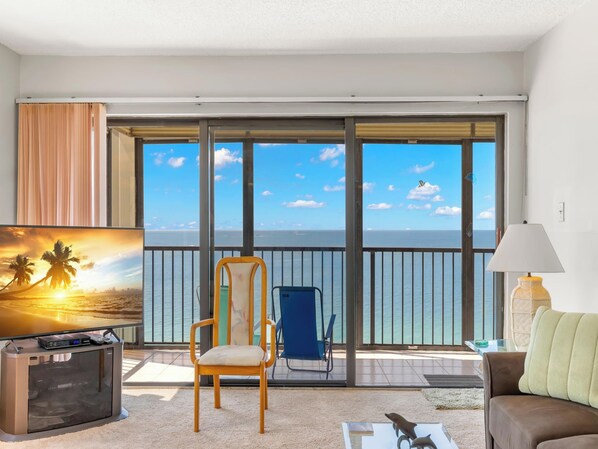 Un salón espacioso con un balcón grande con vistas al mar, un sofá cómodo, un televisor y una silla.