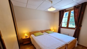 Un dormitorio con dos camas de madera, una ventana con cortinas, una lámpara de techo y una mesita de noche con lámpara.