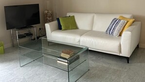 Un salon moderne avec un canapé en cuir blanc, une table basse en verre, un téléviseur à écran plat et un tableau accroché au mur.