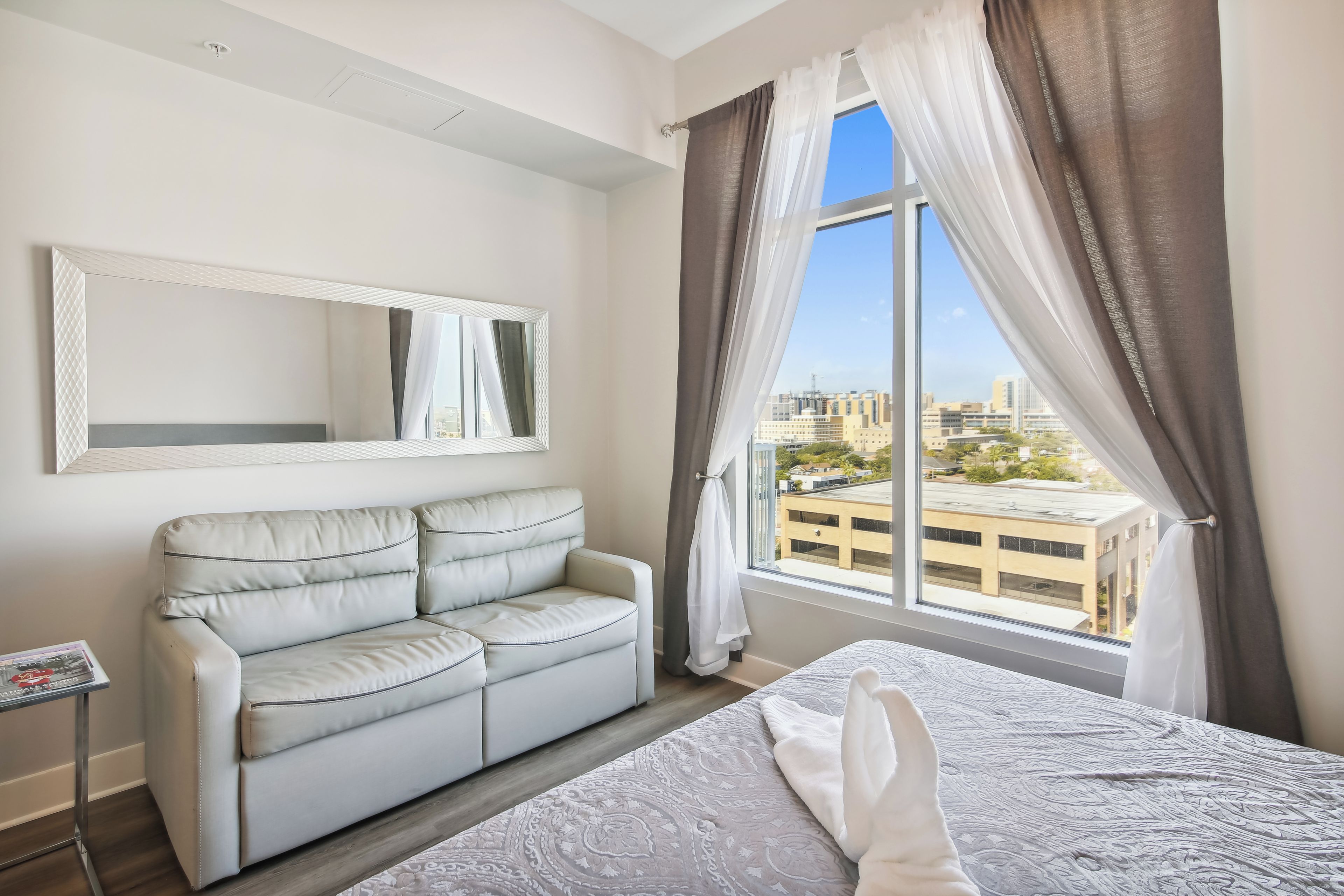 A hotel room with a bed, a sofa, a window with curtains, and a city view.