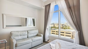 A hotel room with a bed, a sofa, a window with curtains, and a city view. - Emerald 705 by Ryson Vacation Rentals (Galveston)