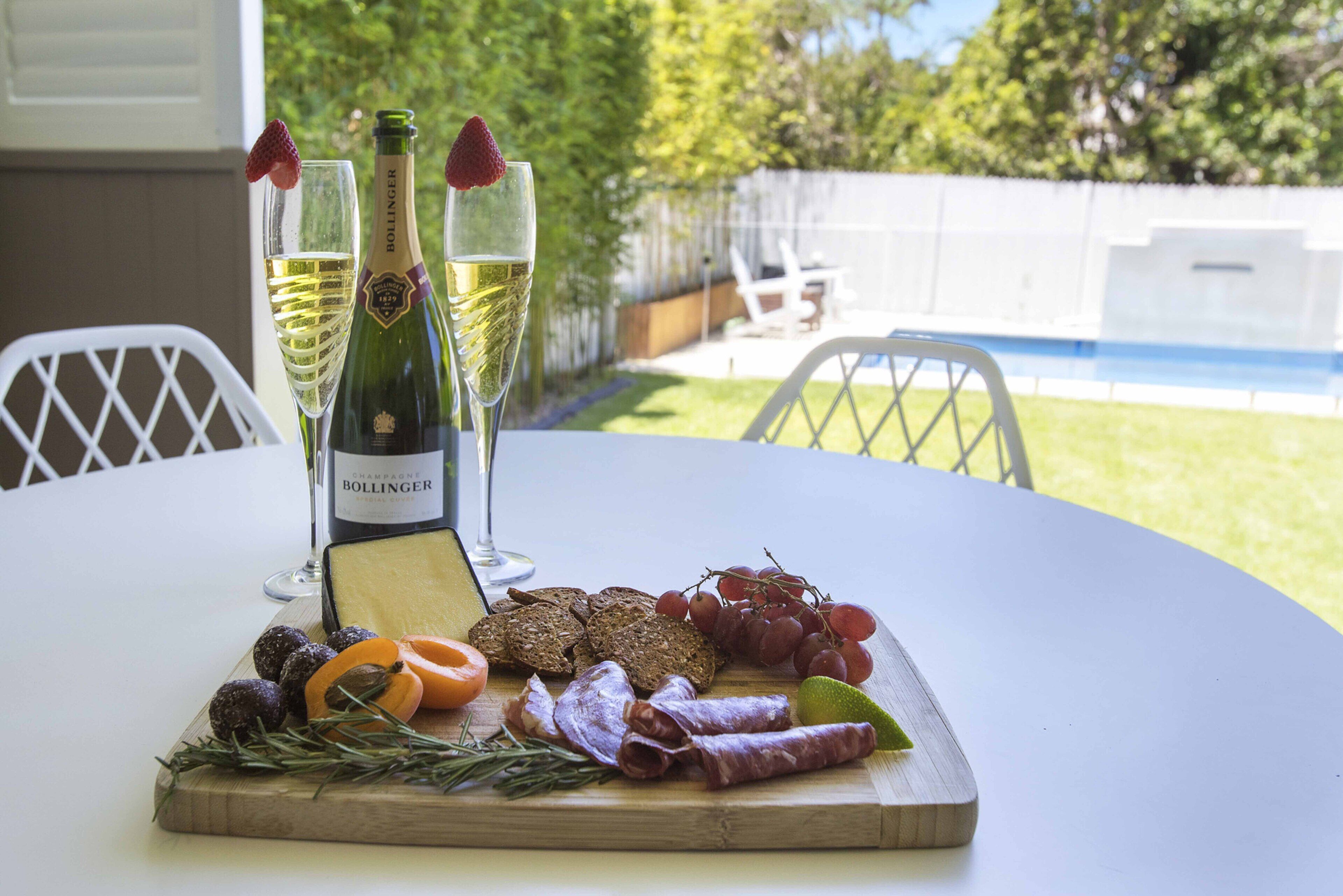A table set with a bottle of Bollinger champagne, two glasses, a cheese board with various accompaniments, and a view of a backyard with a pool.