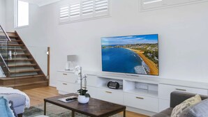 A modern living room with a large flat-screen TV mounted on the wall, a wooden staircase, a white console table with a vase of flowers, and a grey sofa. - Manly Oasis, 5bd, 3 King bd,Pool (Manly)