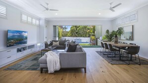 A spacious living room with a large flat-screen TV, a grey sofa, a dining table with chairs, and a view of a backyard with a pool. - Manly Oasis, 5bd, 3 King bd,Pool (Manly)