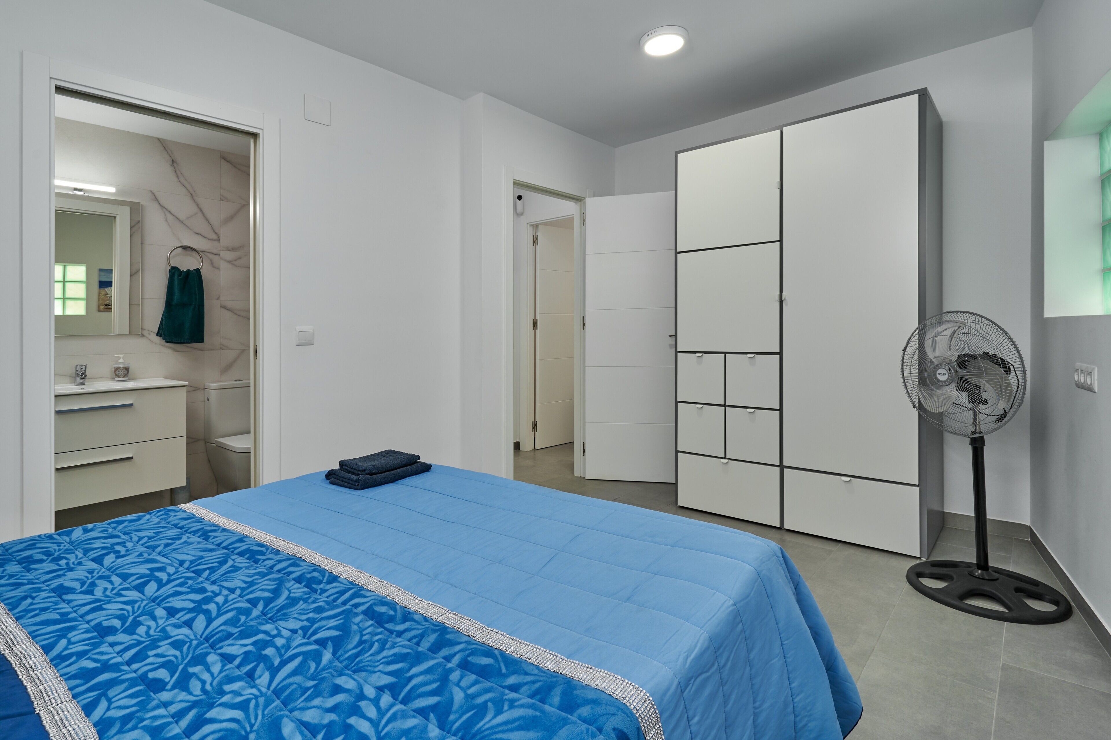 Un dormitorio con cama, armario, espejo, ventilador y baño a la vista a través de una puerta abierta.