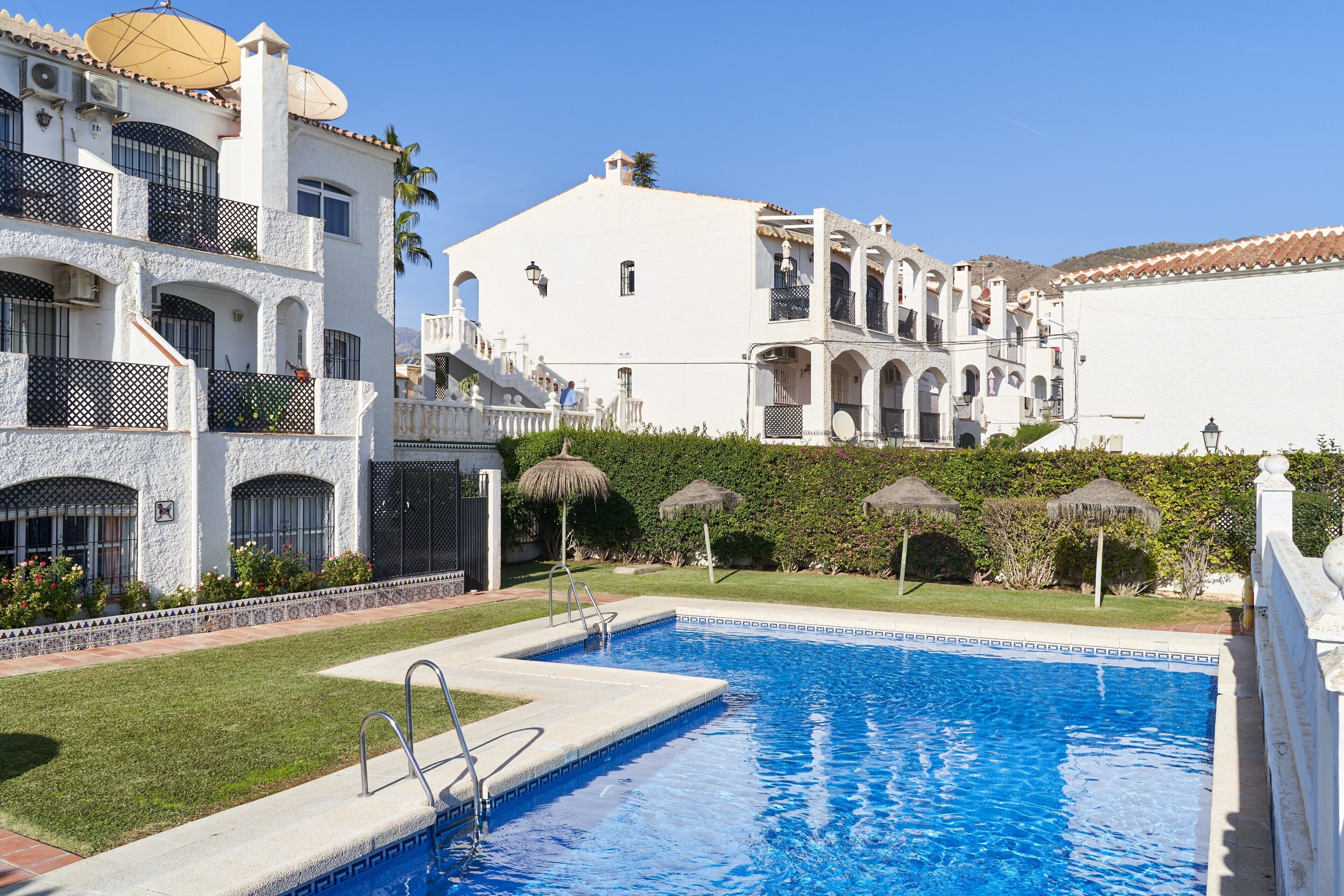 A swimming pool with a stone border, a white building with balconies, and a clear blue sky.