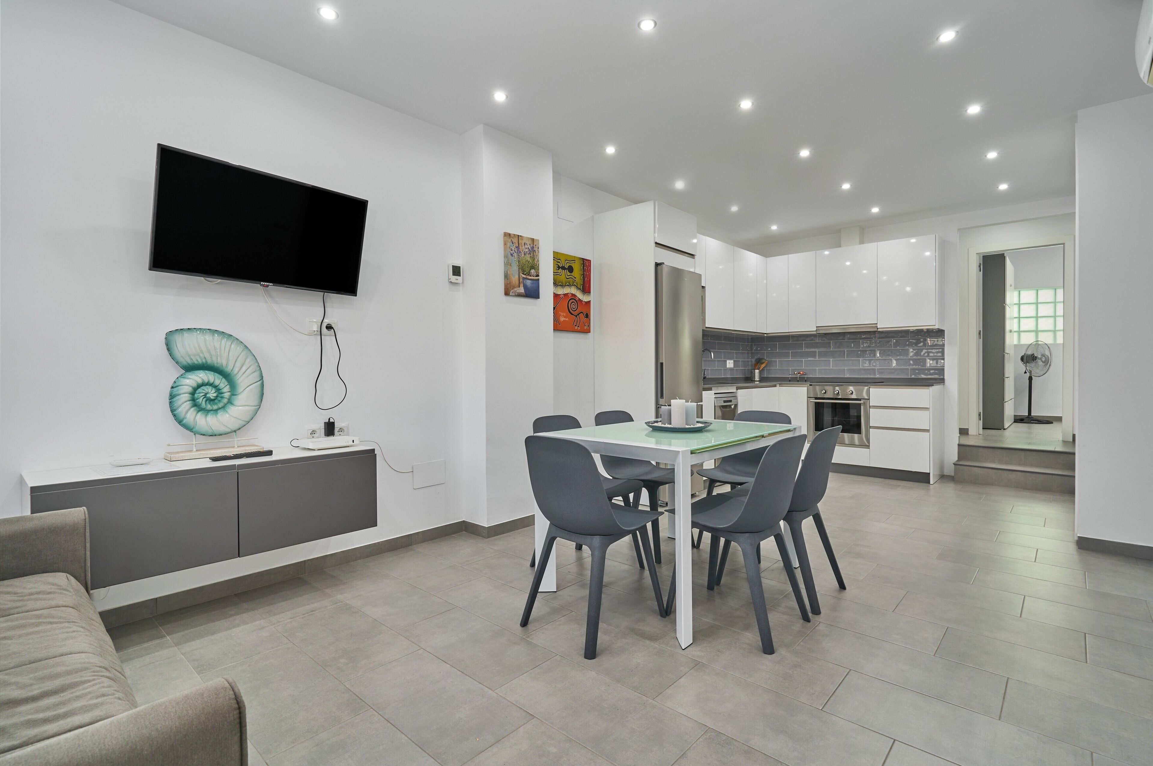A modern kitchen with white cabinets, a stainless steel refrigerator, and a flat-screen TV mounted on the wall.