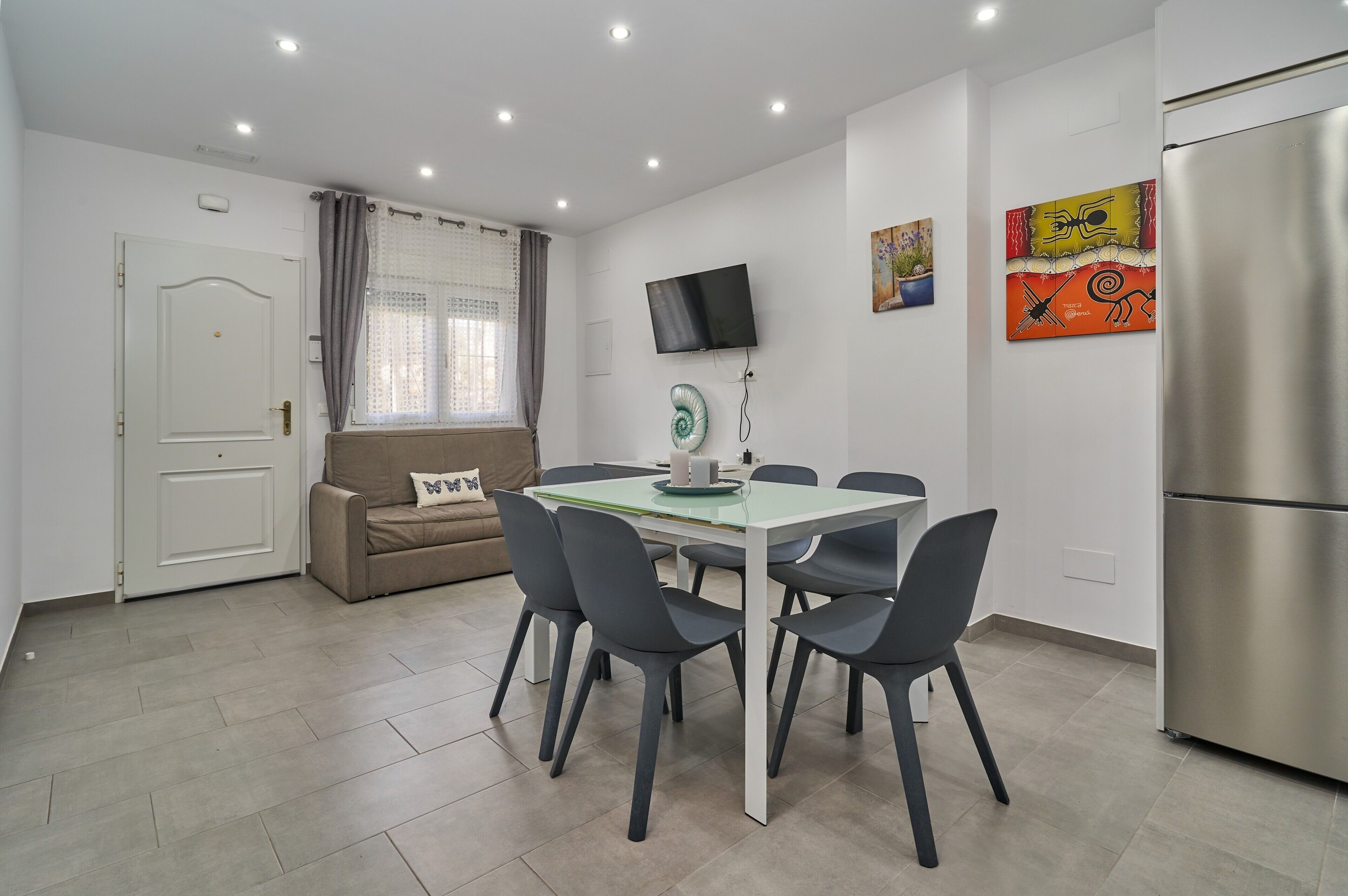 A modern kitchen with a stainless steel refrigerator, a dining table with chairs, a sofa, a TV mounted on the wall, and a window with curtains.