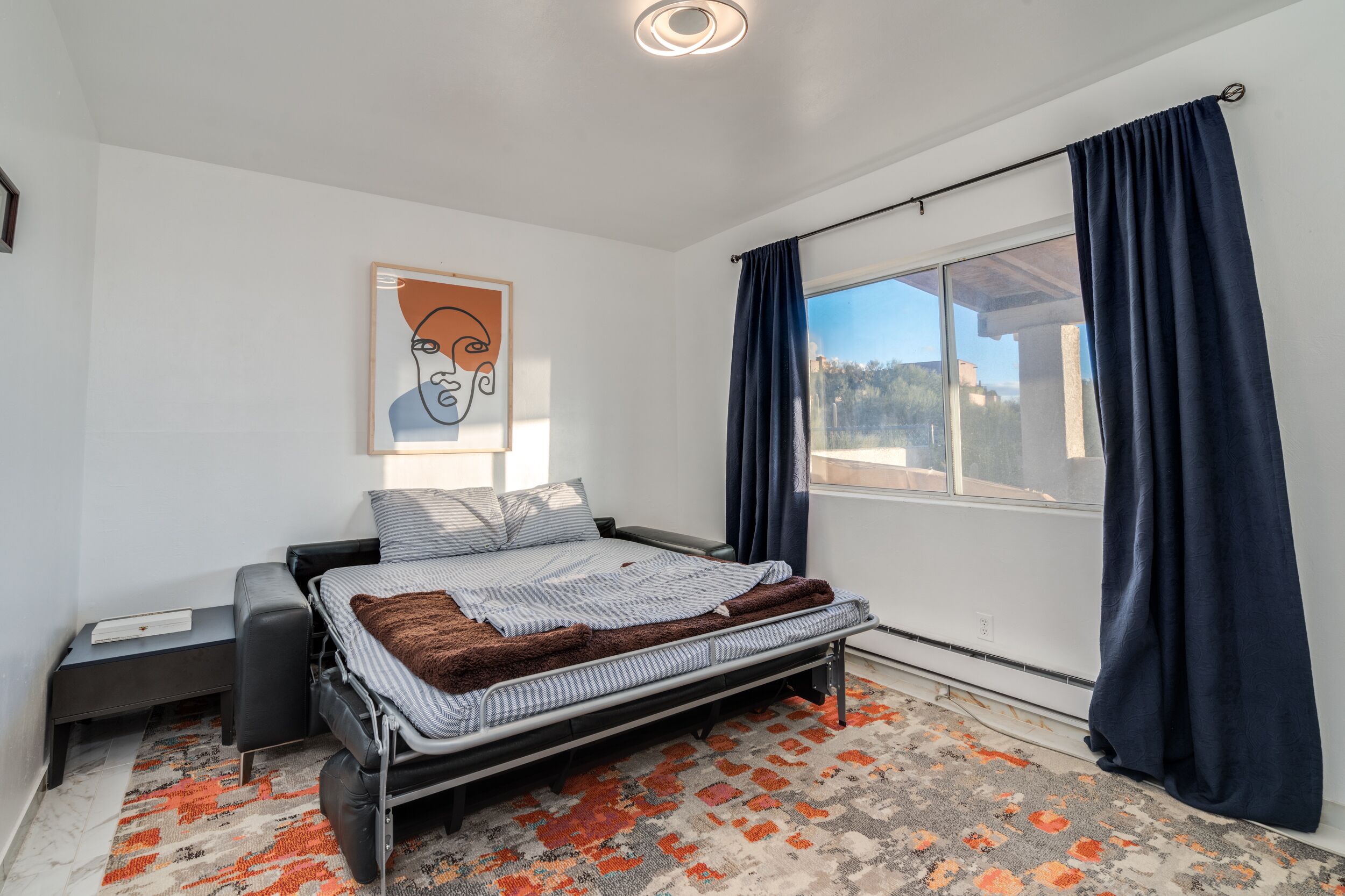 A modern bedroom with a bed, a nightstand, a window with curtains, and a painting on the wall.