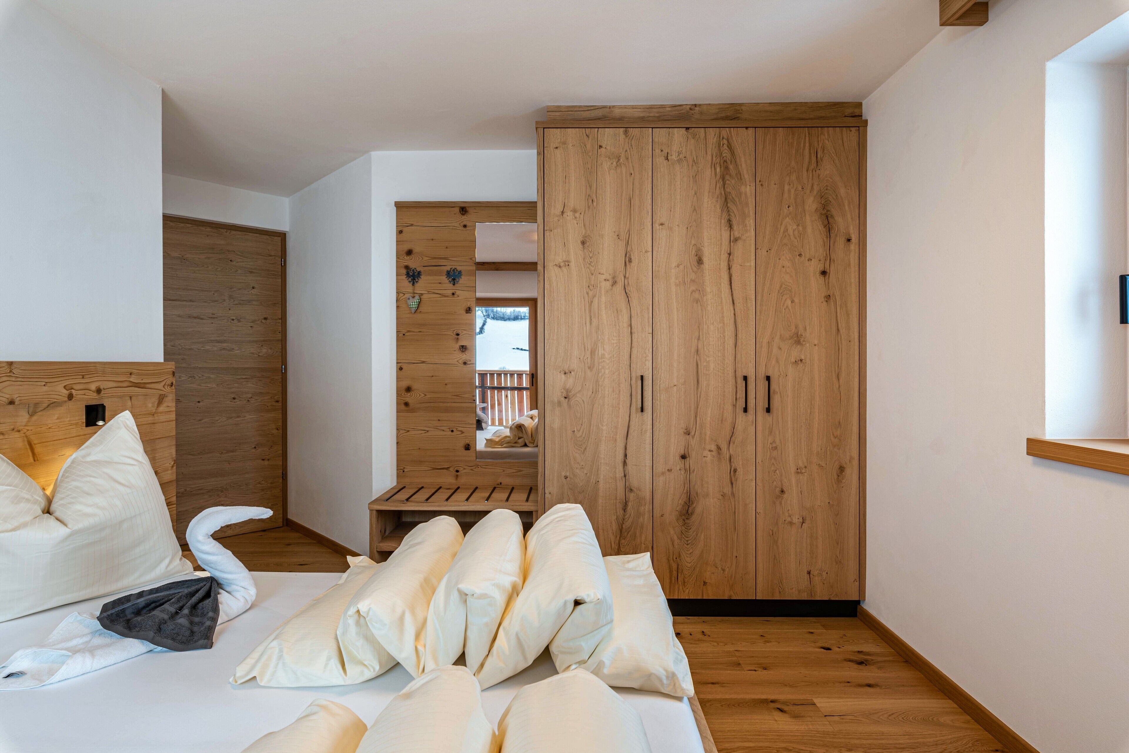 A bedroom with a wooden wardrobe, a bed with pillows, and a small wooden bench.