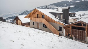 A modern wooden chalet with a steep roof, a chimney, and a view of snow-covered mountains. - Farm 'Enzian' with Mountain View, Shared Terrace and Wi-Fi (Funes)
