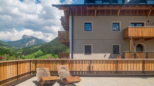 A multi-story building with balconies, a wooden deck with lounge chairs, and a mountainous landscape in the background.