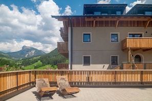 Un immeuble à plusieurs étages doté de balcons, une terrasse en bois avec des chaises longues, et un paysage montagneux en arrière-plan.
