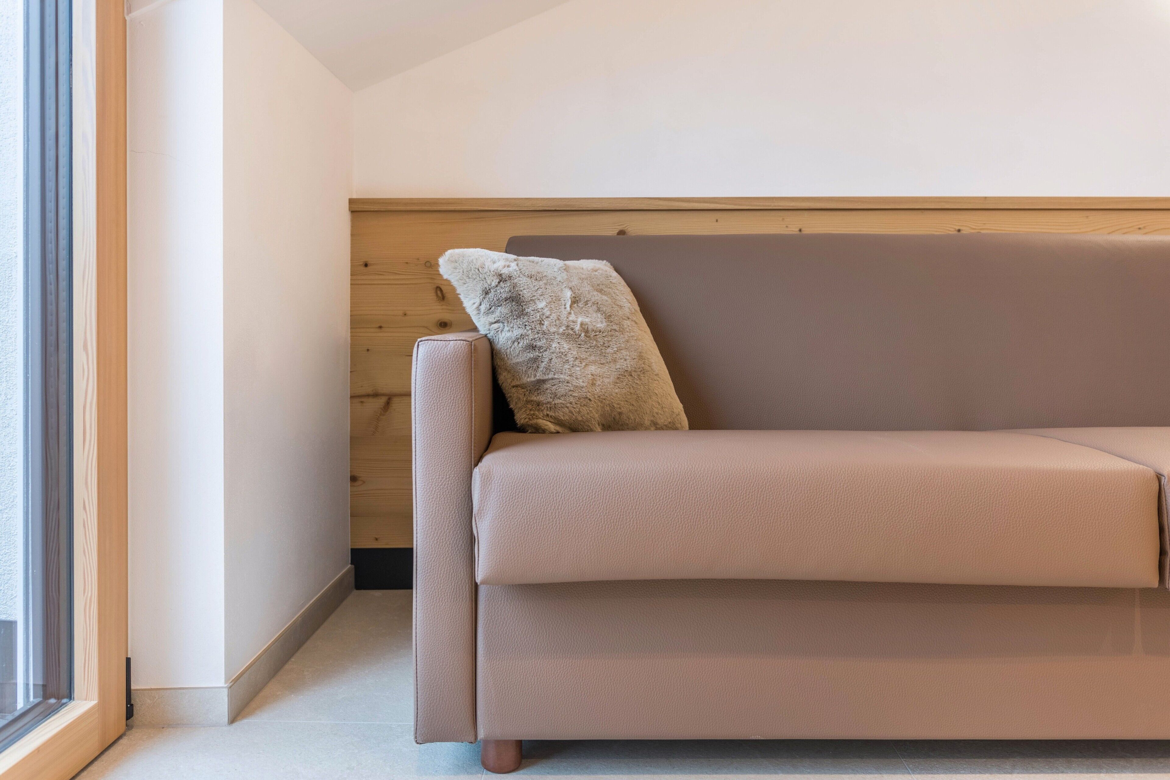 Ein beiges Sofa mit einer strukturierten Kissenlehne an einer Holzwand, ein Fenster links und ein gefliester Boden.