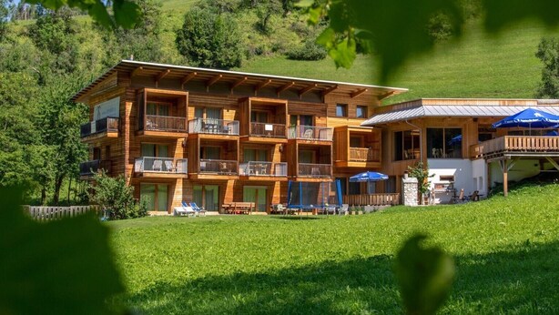 A multi-story wooden building with balconies, a covered outdoor seating area, and a grassy foreground. - Apartment "Le Table C" with Mountain View, Garden & Wi-Fi (St. Vigil in Enneberg)