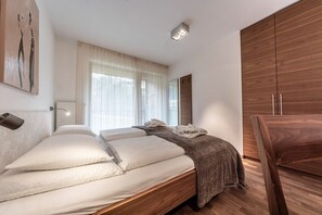 A bedroom with a bed, a wooden wardrobe, a bedside table, and a window with sheer curtains.