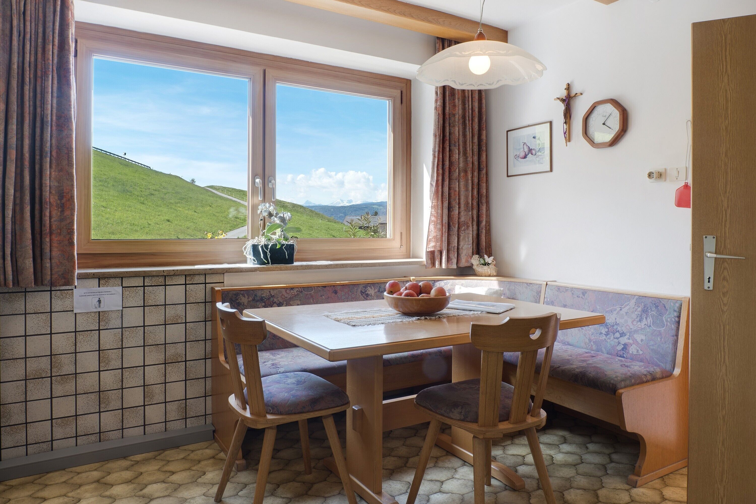 Een eethoek met een tafel, stoelen, een bank, een raam met uitzicht op een groen landschap, een hanglamp en een klok aan de muur.
