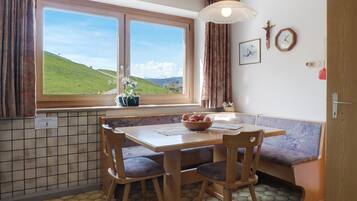 Een eethoek met een tafel, stoelen, een bank, een raam met uitzicht op een groen landschap, een hanglamp en een klok aan de muur.