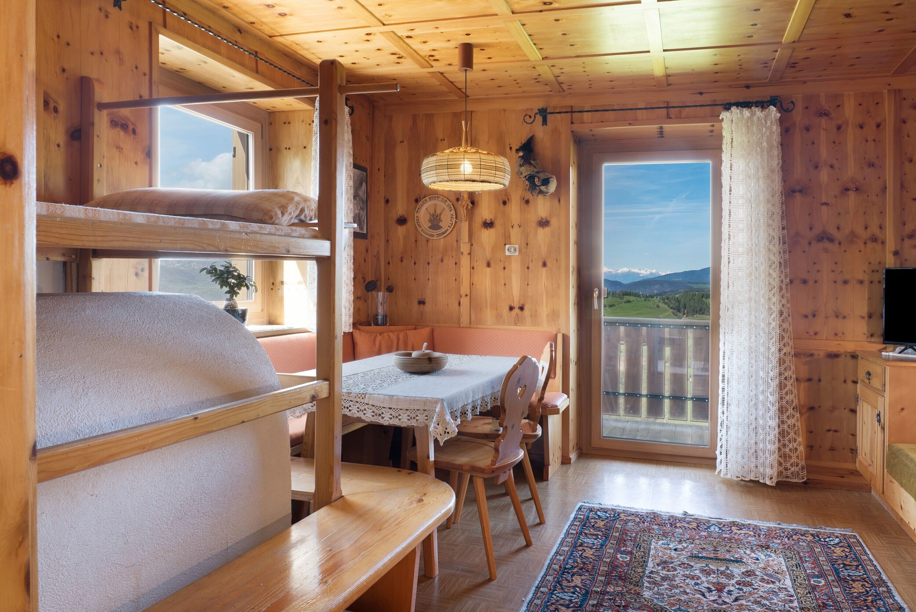 A wooden cabin with bunk beds, a dining table, chairs, a rug, a television, and a view of the outdoors.