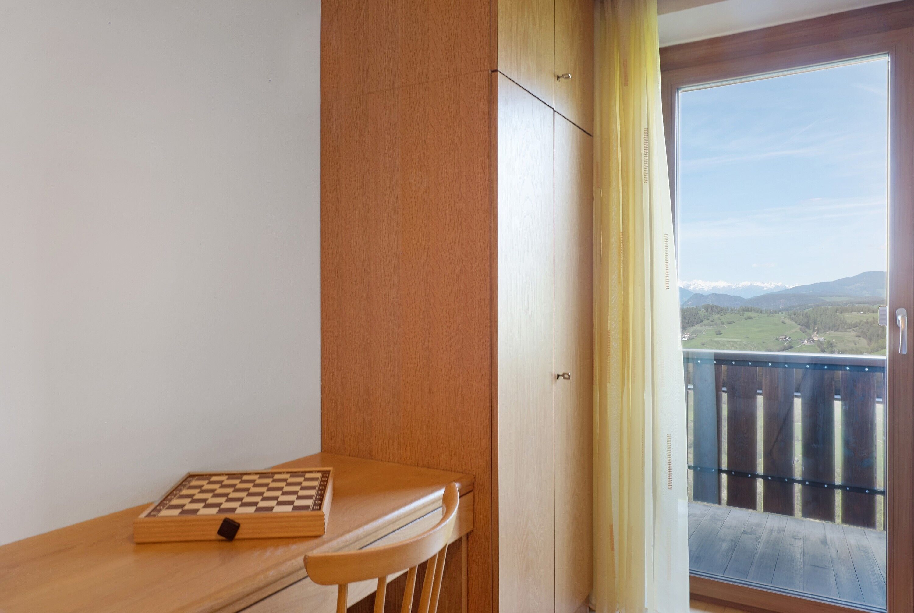 Ein Zimmer mit einem Holztisch, einem Schachbrett und Blick auf einen Balkon mit Geländer.