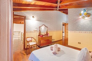 Une chambre avec un plafond en bois, un lit recouvert d’une courtepointe blanche, une commode en bois, une chaise et un mur orné d’un luminaire sur le thème de Pokémon.