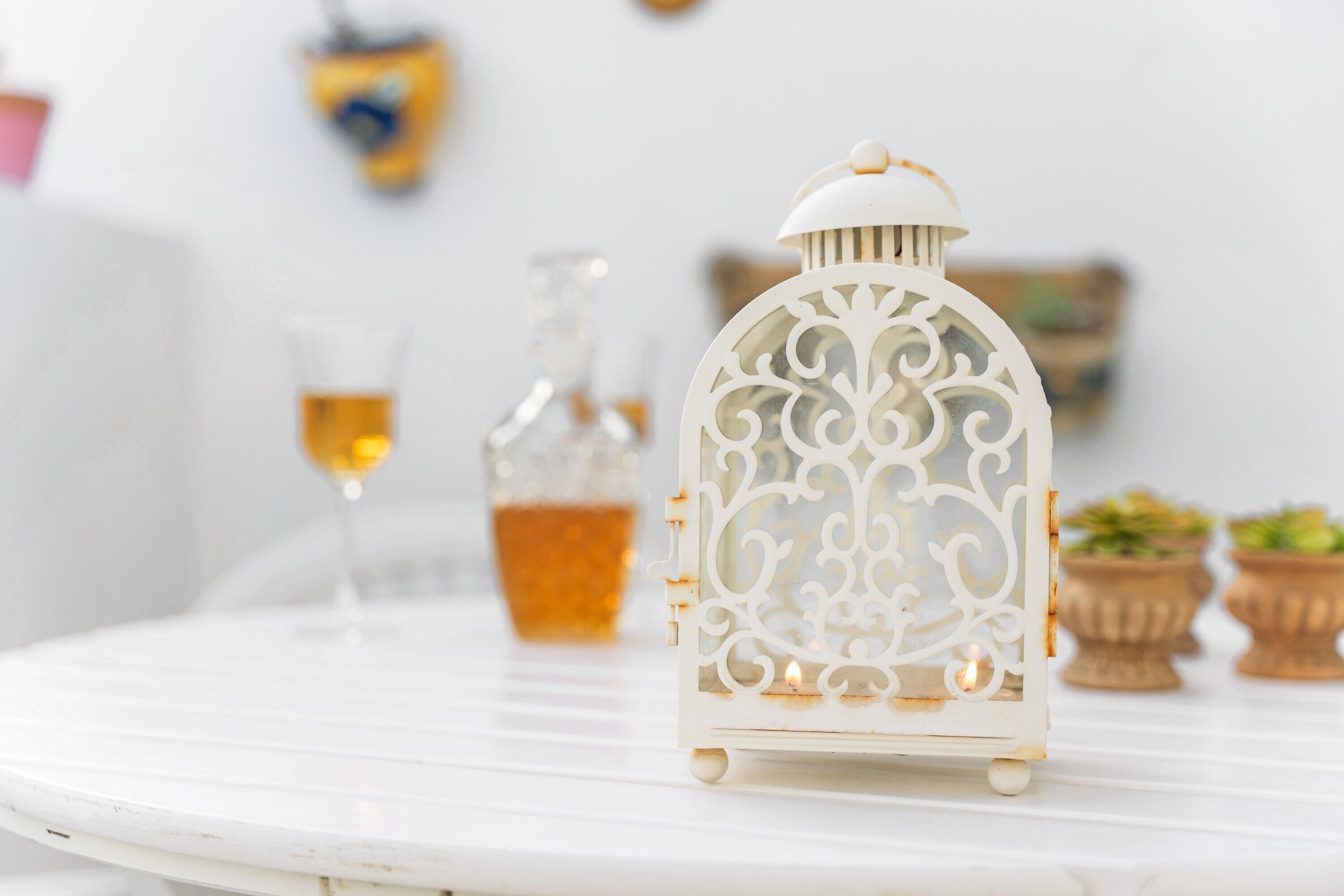 Een witte tafel met een decoratieve lantaarn, een glazen fles en een glas amberkleurige vloeistof.