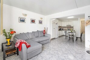 A living room with a grey sofa, a dining table with chairs, a kitchen area, and a small side table with a vase of flowers.