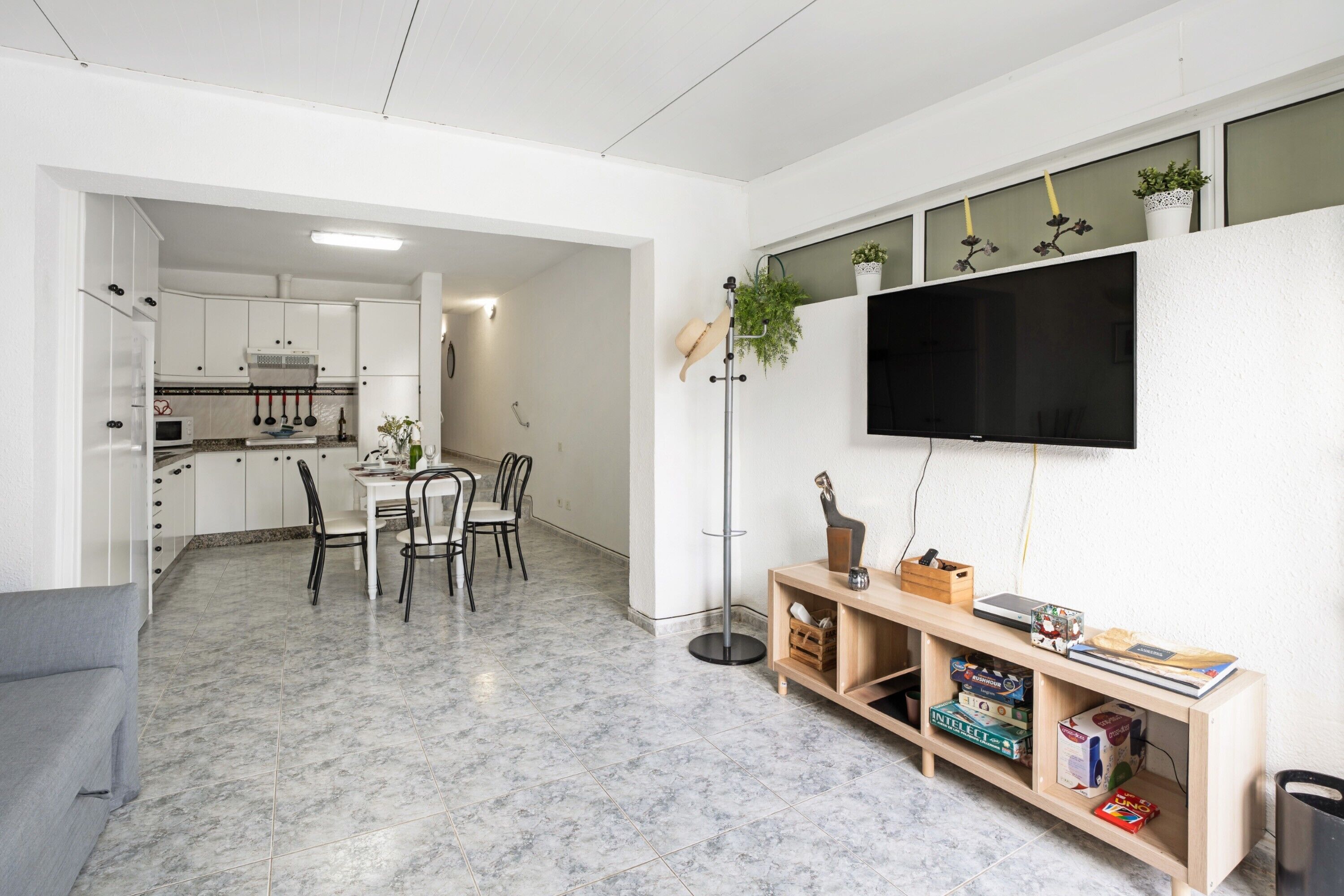 A modern kitchen with white cabinets, a dining area with chairs, a TV mounted on the wall, and a bookshelf with books and decorative items.