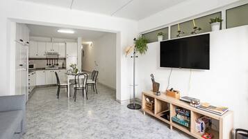 A modern kitchen with white cabinets, a dining area with chairs, a TV mounted on the wall, and a bookshelf with books and decorative items.