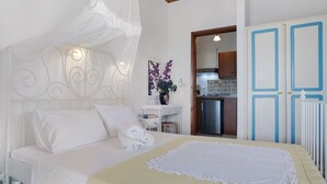 A neatly made bed with white linens, a bedside table with a vase of purple flowers, and a view into the adjacent kitchen area. - Studio Apartment "Areti Orfeas Studio" near Beach with Balcony & Wi-Fi (Zakynthos)