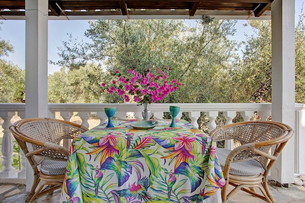 Un espace extérieur abrité, aménagé avec des tables recouvertes d’une nappe à motifs floraux, entouré de chaises en osier, et agrémenté d’un vase rempli de fleurs roses.