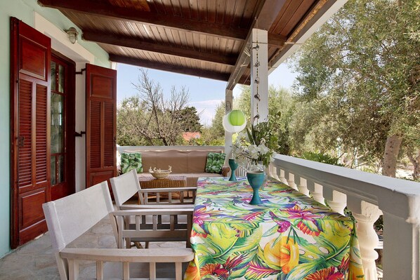A covered outdoor seating area with a table and chairs, a wooden bench, and a view of trees and buildings.