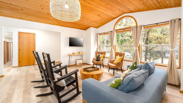 Uma sala de estar espaçosa com teto de madeira, uma janela grande com cortinas, um sofá azul e uma televisão na parede.