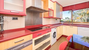 Cocina compacta con horno integrado, microondas y lavavajillas, una pequeña zona de comedor con mesa y sillas, y ventana con vistas a palmeras.