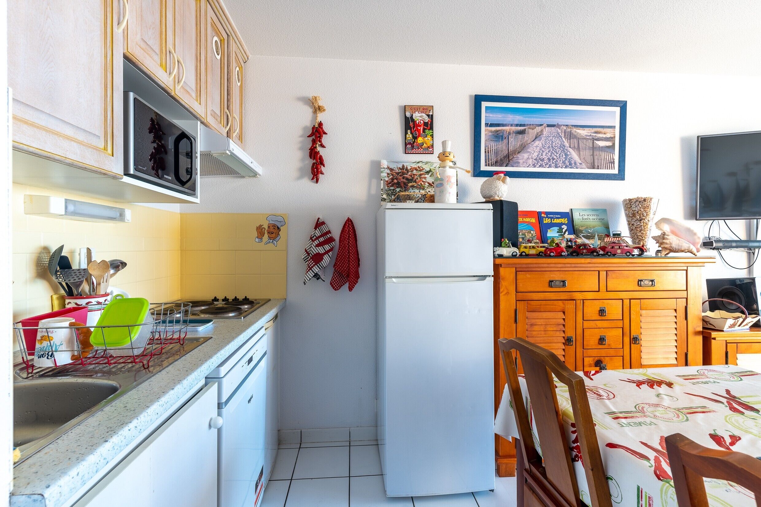Una cucina con frigorifero, piano cottura e microonde. Sono presenti armadietti in legno, un tavolo da pranzo con sedie e una televisione.
